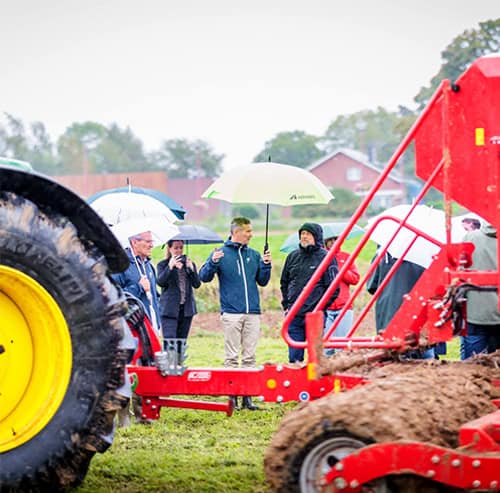 Several people are standing with umbrellas on a wet field, watching agricultural machinery at work.
